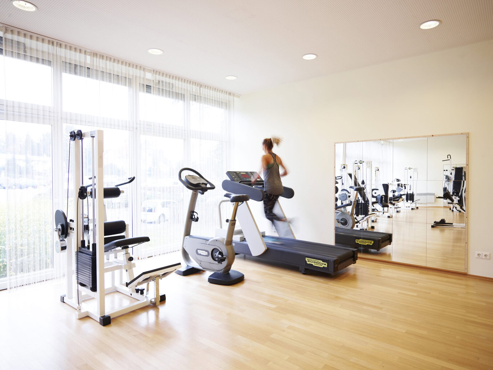 Zu sehen ist ein lichtdurchfluteter Fitnessraum. Es gibt zahlreiche, moderne Geräte für Kraftsport und Cardio.
