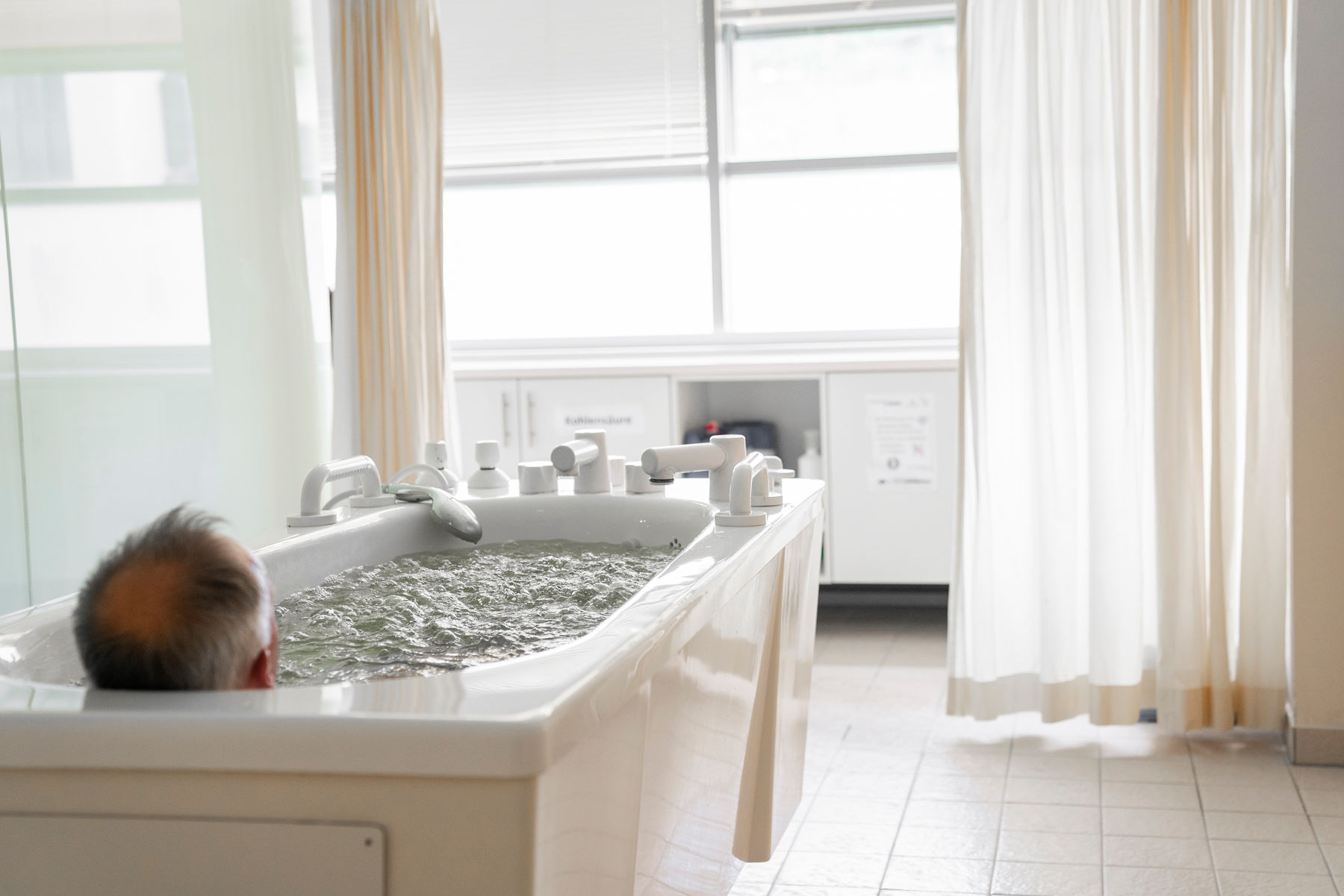 Ein Mann nimmt ein Bad. Er liegt in einer Badewanne in einem lichtdurchfluteten Raum.