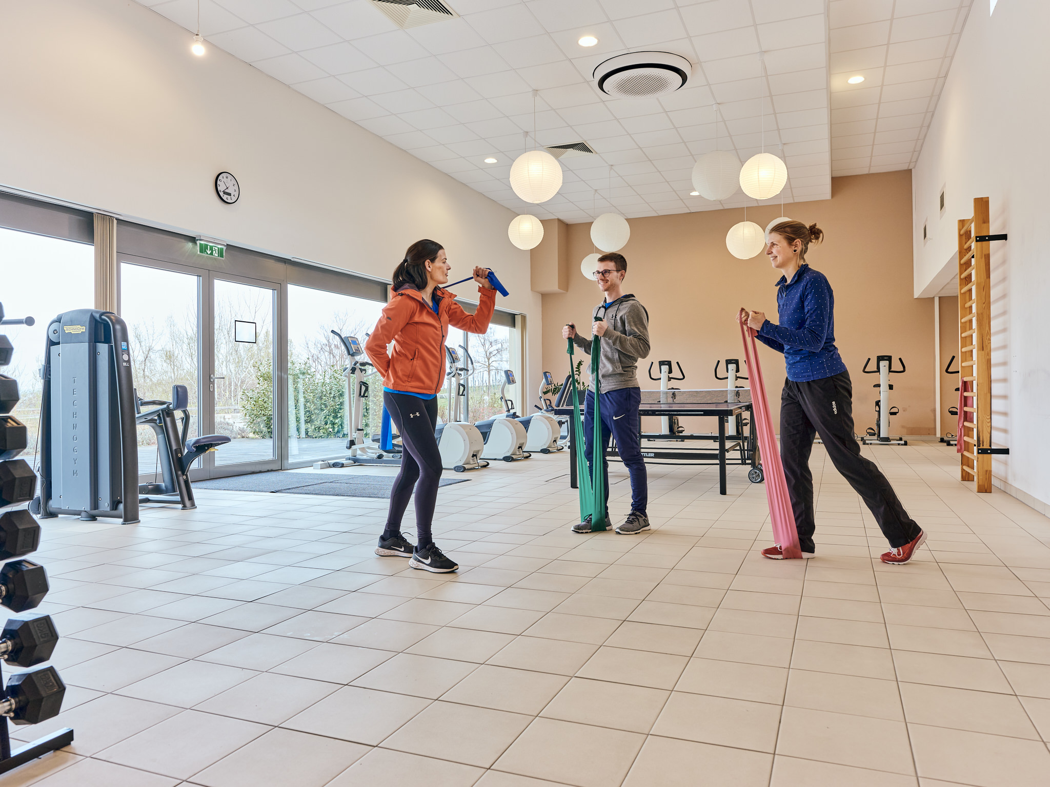 Eine Trainerin übt mit einer Frau und einem Mann mit Therabändern in einem Fitnessraum.