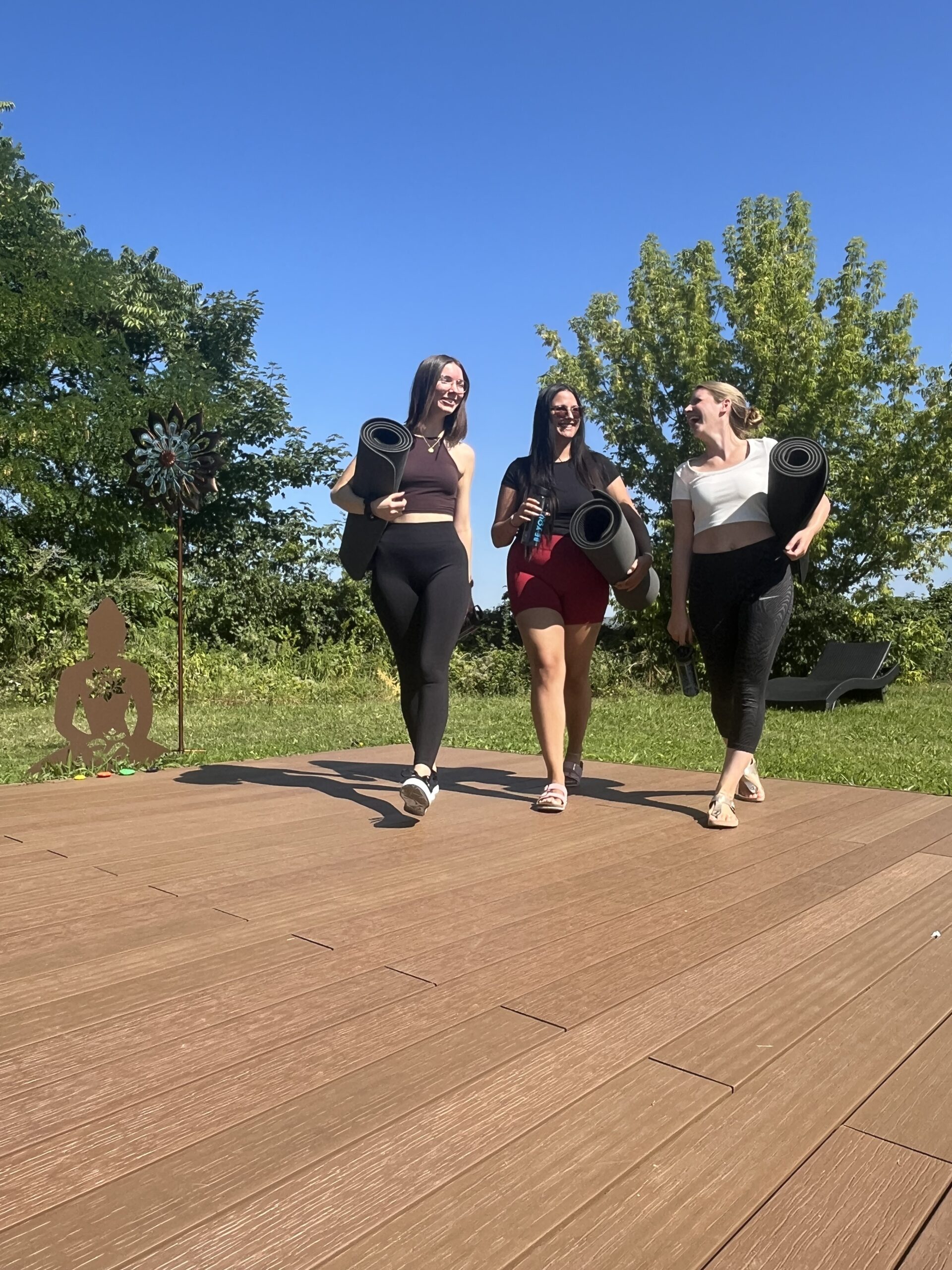 Drei junge Frauen in Yogakleidung unterhalten sich und tragen ihre Yogamatten. Sie befinden sich auf einem Holzdeck und hinter ihnen sind Bäume zu sehen.
